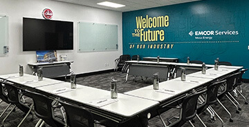 Several desks places in a U shape facing the front of the room with a tv on the wall with the words Welcome to the Future - EMCOR Services on the side wall. 