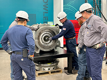 Four people wearing safety helmets inspecting or working on a large industrial machine component in a workshop or training facility.