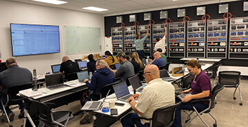 A group of people working on laptops in a control room or training lab, facing a large display screen and a wall with numerous electrical or network panels.